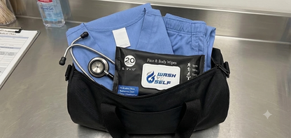 A black medical-style duffel bag on a stainless steel counter holding folded blue scrubs, a stethoscope, and a packet of Wash & Self face and body wipes.